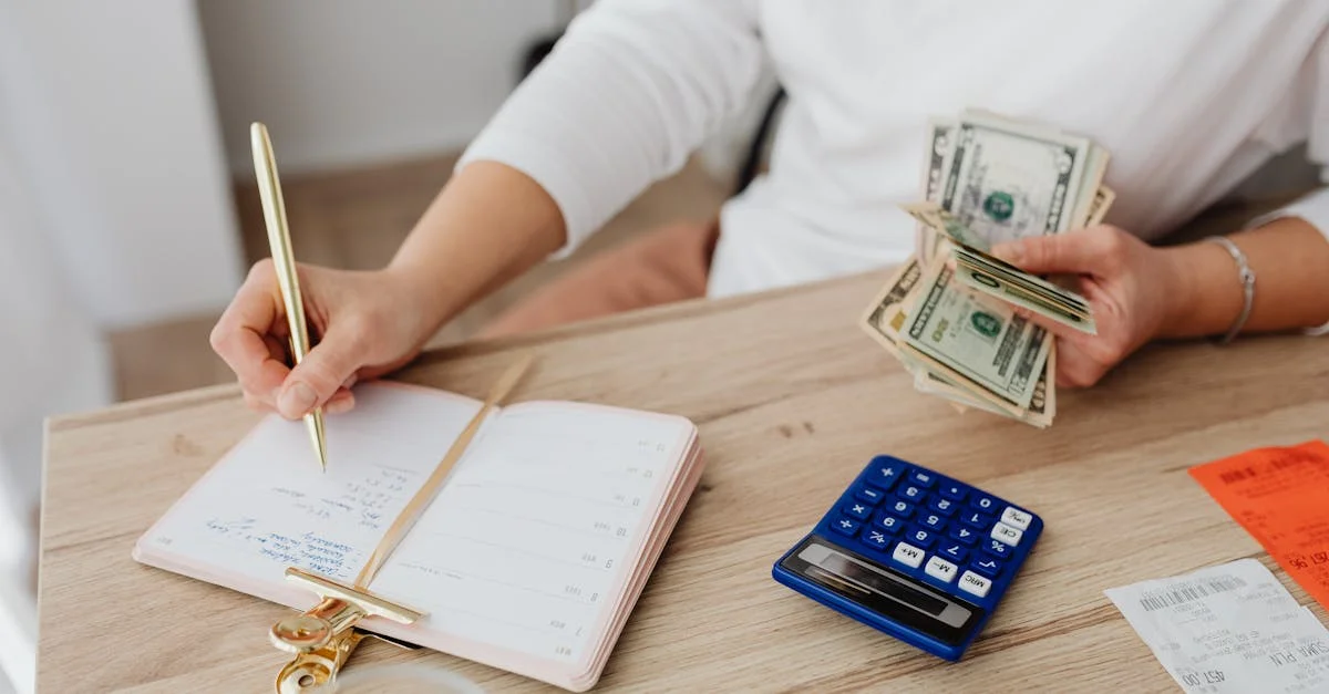 Adult holding cash and writing in planner while using a calculator at home.