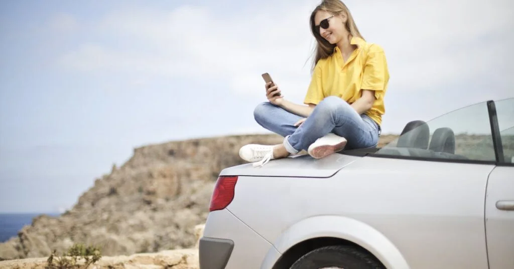 Smiling young woman sitting on a car at the beach in Mgarr, Malta using a smartphone.